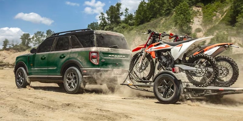 Un vehículo Ford Bronco remolca un remolque con dos motos de cross en un camino de tierra.