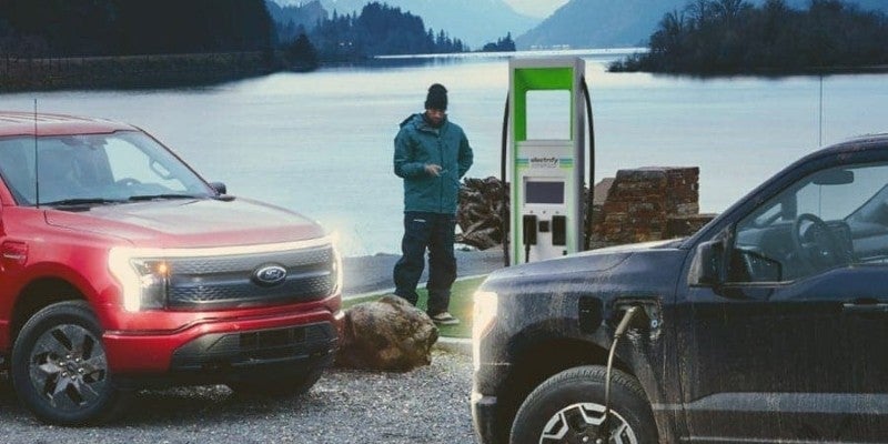 Dos camionetas pickup?F-150?Lightning estacionadas frente a frente con los faros encendidos y un hombre junto a la estación de carga.