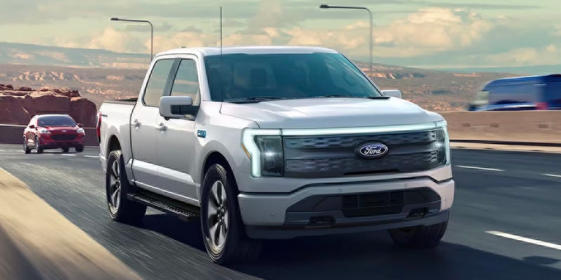 Una camioneta pickup F-150 Lightning de Ford vista desde el lado derecho en marcha, con los faros encendidos.