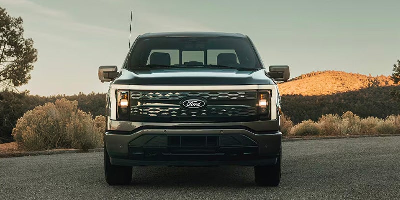 Una camioneta pickup?Ford F-150 Lightning aparcada vista desde el frente con los faros encendidos y un paisaje natural impresionante de fondo.
